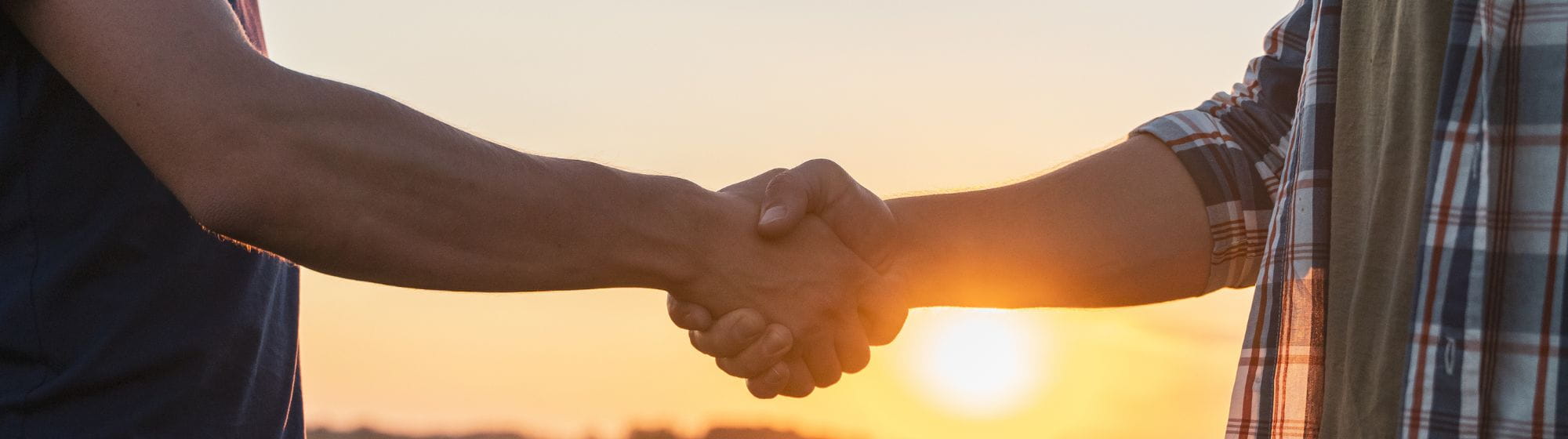Poignée de main entre deux personnes dans un champ agricole au coucher du soleil, symbolisant un accord ou un partenariat.