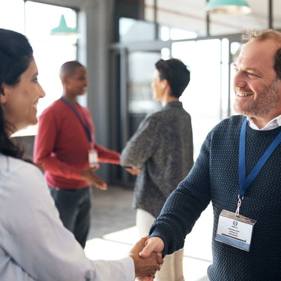 Two smiling people shaking hands in a professional setting, wearing name badges, with other professionals in the background.